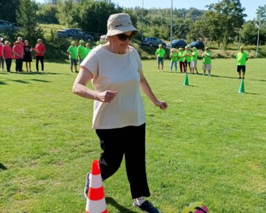 Zbliżenie. Seniorka w kapeluszu i okularach biegnie koło palika. Przed nią piłka. W tle grupy zawodników ubrana na kolorowo.