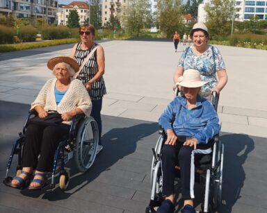 Zbliżenie. Plac. Dwie kobiety pchają wózki, na których siedzą seniorki. W tle zieleń niska, krzewy, drzewa i bloki.