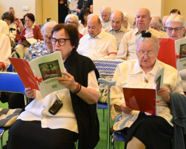 Sala. Grupa seniorów siedzi na sali i trzyma w ręku śpiewniki. Seniorzy ubrani są w jasne koszule i ciemny dół.