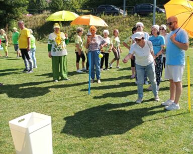 Boisko. Grupa seniorów. Kobieta na odległość wrzuca do kosza niebieskie piłeczki. Część seniorek trzyma kolorowe parasole.