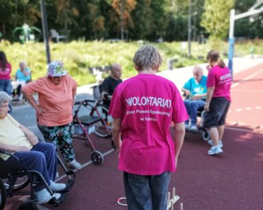 Boisko. Grupa seniorów wykonuje ćwiczenia. Na boisku dwoje wolontariuszy w różowych podkoszulkach z napisem 