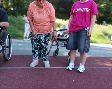 Boisko. Młoda wolontariuszka stoi z seniorką. Seniorka ma na głowie kapelusz. Przed nimi pudełko ze szmatką na brzegu.