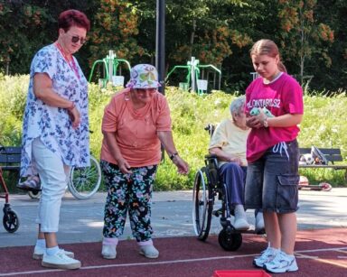 Boisko. Cztery osoby. Dwie wolontariuszki i seniorka wrzucają do pudełka woreczki w kropki. W tle seniorka na wózku, ławki.