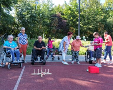 Boisko. Grupa seniorów i kilku wolontariuszy grają w gry zręcznościowe. W tle trawiasta skarpa i fit park.