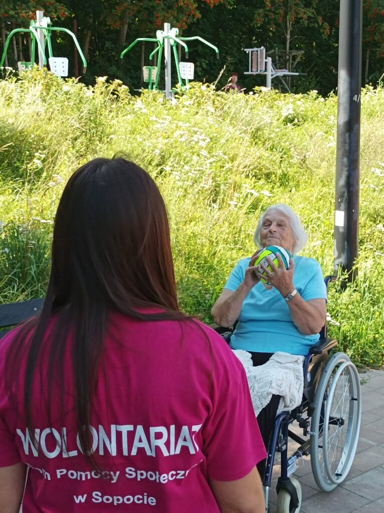 Zbliżenie. Wolontariuszka i seniorka na boisku. Seniorka trzyma w rękach piłkę. W tle trawiasta skarpa. Fit Park.