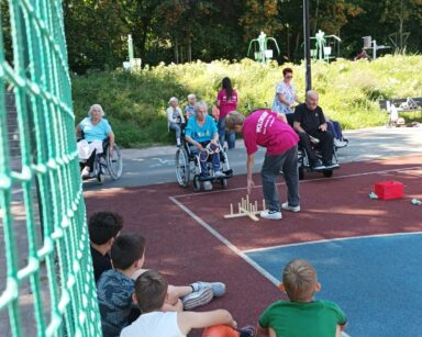 Boisko. Grupa seniorów i kilku wolontariuszy grają w gry zręcznościowe. W tle trawiasta skarpa i fit park.