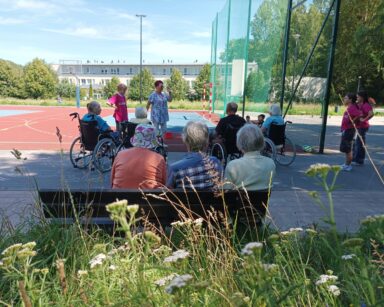 Boisko. Grupa seniorów i kilku wolontariuszy grają w gry zręcznościowe. W tle trawiasta skarpa i fit park.