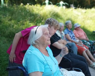 Zbliżenie. Grupa seniorów na wózku siedzi na boisku. Obok jednej z seniorek stoi młody wolontariusz. W tle trawiasta skarpa.