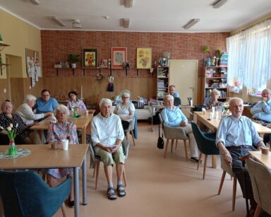 Sala. Przy stołach siedzi grupa seniorów. W tle na ścianach obrazy w słoneczniki, książki na półkach. Kanapa i poduszki.