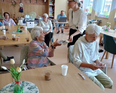 Sala. Kobieta w jasnej bluzce i ciemnych spodniach stoi przy seniorach, którzy siedzą przy stołach i rozdaje im ulotki.