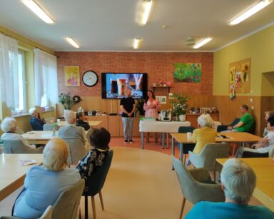 Sala. Para stoi przy stole. Na nim szklanki i sprzęt do kawy. W tle na monitorze dwie kawy. Przy stołach siedzą seniorzy.