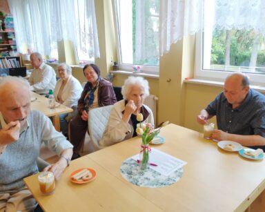 Zbliżenie. Trzech seniorów przy stoliku degustuje mrożoną kawę. Jedzą łyżeczką z kawy bitą śmietanę. W tle grupa seniorów.