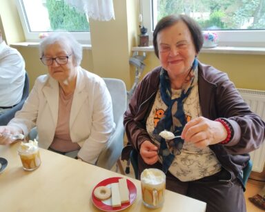 Zbliżenie. Przy stole siedzą dwie seniorki. Obie panie trzymają w ręku łyżeczkę z bitą śmietaną. Na stole mrożona kawa.