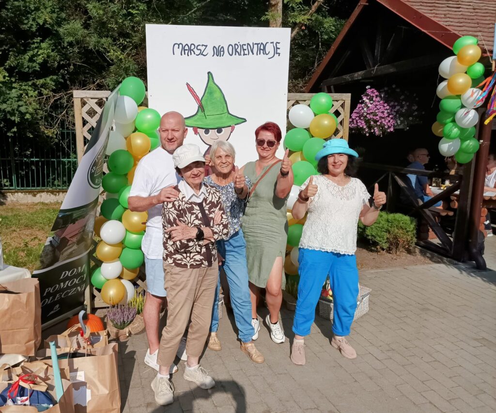 Zbliżenie. Cztery kobiety i mężczyzna pozują do zdjęcia. W tle kolorowe balony. Plakat z napisem "Marsz na orientację".