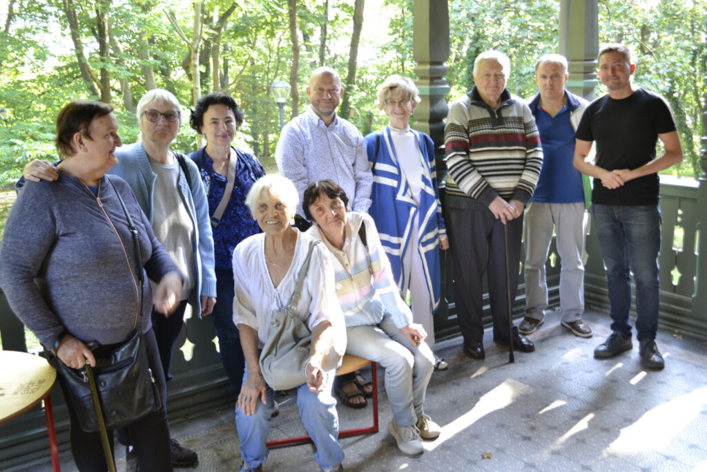 Zbliżenie. Grupa seniorów pozuje do zdjęcia grupowego. Seniorzy stoją na werandzie przy drewnianej, rzeźbionej balustradzie.