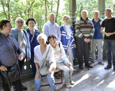 Zbliżenie. Grupa seniorów pozuje do zdjęcia grupowego. Seniorzy stoją na werandzie przy drewnianej, rzeźbionej balustradzie.