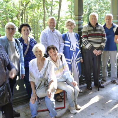 Zbliżenie. Grupa seniorów pozuje do zdjęcia grupowego. Seniorzy stoją na werandzie przy drewnianej, rzeźbionej balustradzie.