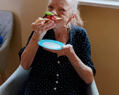 Zbliżenie. Seniorka w ciemnej bluzce w białe kropki siedzi na fotelu. Kobieta je bruschettę. W ręku trzyma talerzyk.