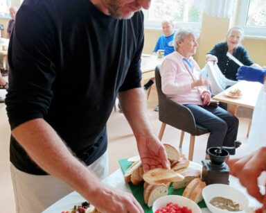 Sala. Mężczyzna w ciemnej bluzce trzyma w ręku bułkę, w drugiej nóż z serkiem. Na stole produkty do zrobienia bruschetty.