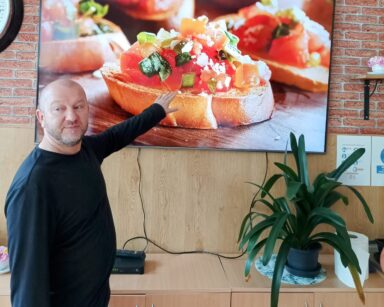 Zbliżenie. Mężczyzna pokazuje na monitor telewizora. Na ekranie bruschetta, czyli bułka z pomidorami, bazylią i serem.