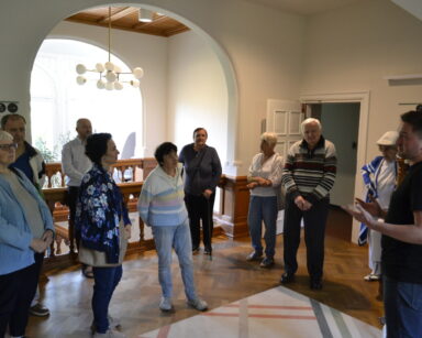 Korytarz. Białe ściany, wiszące żyrandole, piękne drewniane drzwi, przy drewnianych schodach grupa seniorów.