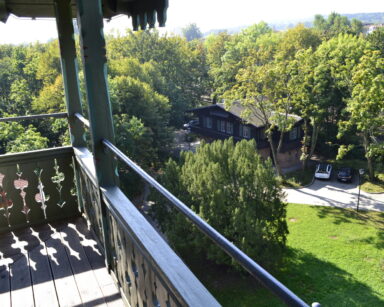 Drewniany balkon, z rzeźbioną balustradą. Z balkonu widok na park i pobliskie zabytkowe domki.