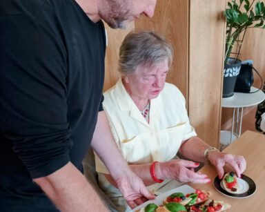 Zbliżenie. Mężczyzna w ciemnej bluzce trzyma talerz z bruschettą, seniorka siedzi przy stole, kładzie przekąskę na talerz.