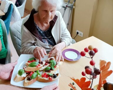 Zbliżenie. Seniorka w siwych włosach i białej bluzie siedzi przy stole. Wybiera z talerza bruschette. Obok leży jej talerzyk.