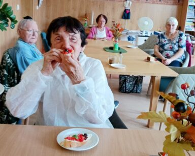 Sala. Seniorka w białym fartuchy siedzi przy stoliku, trzyma przy ustach bruschettę. w tle siedzą seniorzy przy stołach.