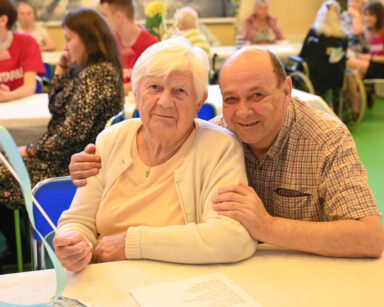 Sala. Para seniorów pozuje do zdjęcia. Seniorka trzyma niebieską wstążkę, senior obejmuje seniorkę za ramiona.