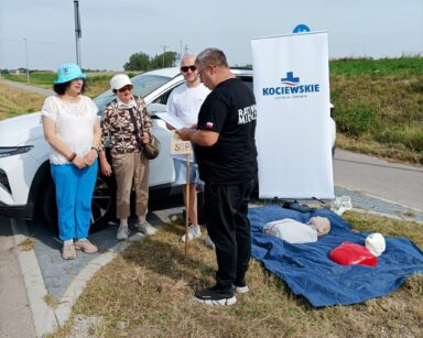 Zbliżenie. Przy drodze stoi ratownik medyczny i trzyma kartkę. Obok dwie seniorki i mężczyzna oraz fantomy do ćwiczeń.