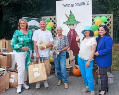 Zbliżenie. Grupa pięciu osób pozuje do wspólnego zdjęcia. Mężczyzna trzyma dylom i papierowe torby. W tle torby z podarkami.