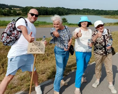 Zbliżenie. Cztery osoby pozują do zdjęcia. Wszyscy unoszą do góry kciuki. Mężczyzna trzyma napis DPS SOPOT. W tle jezioro.