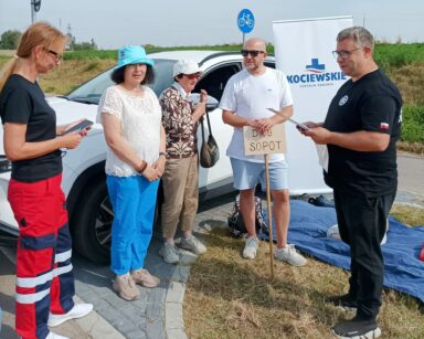 Zbliżenie. Dwóch ratowników medycznych, dwie seniorki i mężczyzna. W tle samochód i napis Kociewskie Centrum Zdrowia.