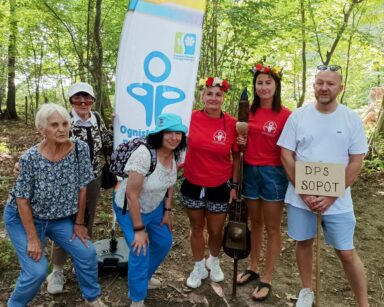 Las. Zbliżenie. Trzy seniorki pozują do zdjęcia z mężczyzną i dwiema kobietami w czerwonych koszulkach i wiankach na głowie.