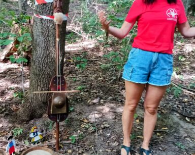 Zbliżenie. Kobieta w czerwonej koszulce stoi w lesie. Przy drzewie oparty jest instrument muzyczny, obok stoi beczka.