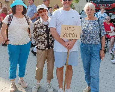 Zbliżenie. Mężczyzna trzyma napis DPS SOPOT, obok stoją trzy seniorki. Wszyscy pozują do zdjęcia. W tle tłum ludzi.