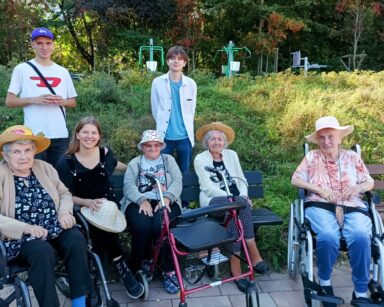 Zbliżenie. Grupa seniorek i trzech młodych wolontariuszy pozują do wspólnego zdjęcia. W tle skarpa porośnięta roślinnością.