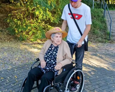 Boisko. Młody mężczyzna pach wózek, na którym jest seniorka. Kobieta ma na głowie kapelusz, mężczyzna czapeczkę z daszkiem.