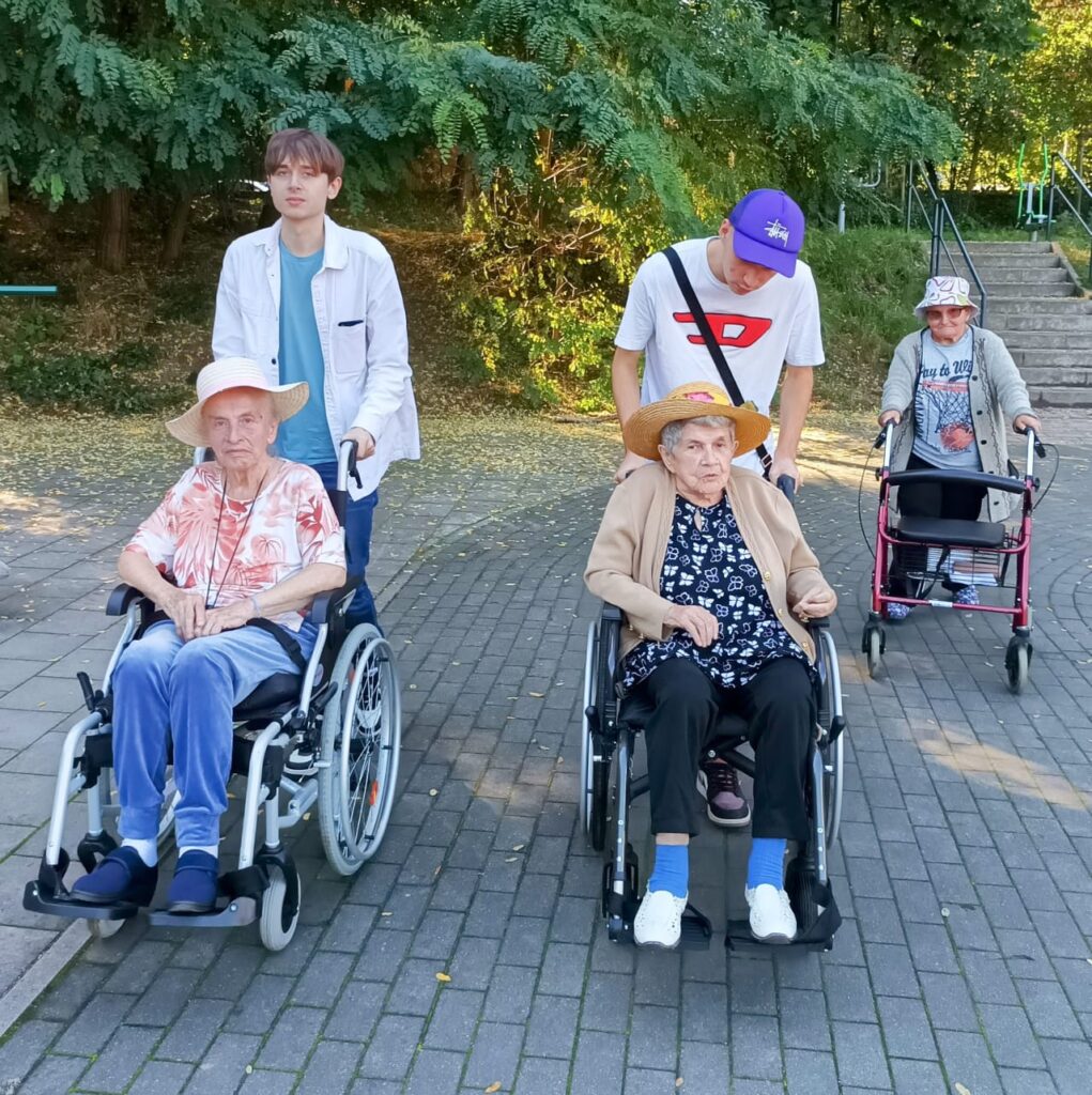 Boisko. Dwóch młodych mężczyzn pcha wózki, na których siedzą seniorki. Za nimi idzie seniorka na chodziku. W tle schody.