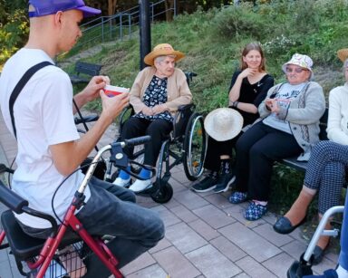 Zbliżenie. Młody mężczyzna oparty o chodzik, trzyma w ręku karteczki. Na ławce siedzą seniorki i młoda kobieta.