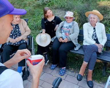 Boisko. Młody mężczyzna siedzi na chodziku. W ręku trzyma karteczki. Przed nim na ławce siedzą seniorki i młoda dziewczyna.