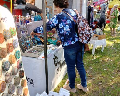 Zbliżenie. Seniorka stoi przy stoisku z biżuterią, kolorowymi pudełeczkami. Kobieta sięga po wisior. W tle balony.