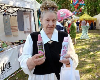 Zbliżenie. Seniorka trzyma w dłoniach dwie butelki z płynem różanym. Obok namiot z obrusami, serwetkami. W tle inne stoiska.