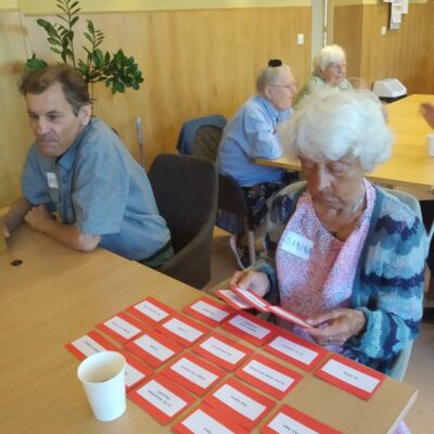 Sala. Stoły. Przy stołach seniorzy. Para przy stole, senior trzyma kubek, seniorka przegląda kartki z napisami.