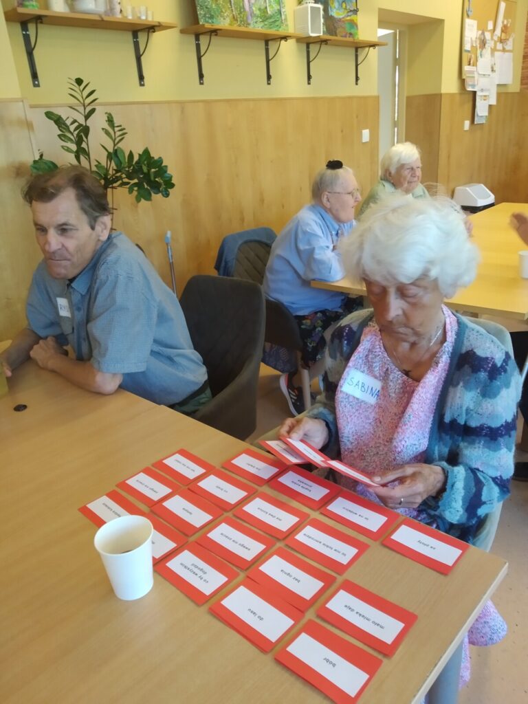 Sala. Stoły. Przy stołach seniorzy. Para przy stole, senior trzyma kubek, seniorka przegląda kartki z napisami.