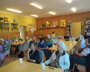 Sala. Seniorzy siedzą przy stołach. Na stołach poustawiane są białe kubeczki.