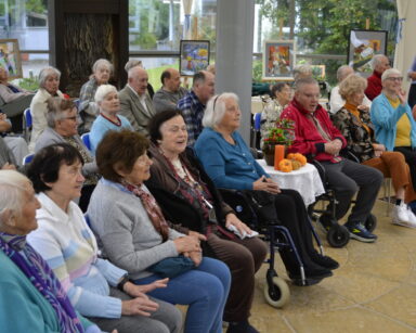 Zbliżenie. Grupa seniorów siedzi w sali i śpiewa piosenki. W tle wystawa obrazów przy oknach.