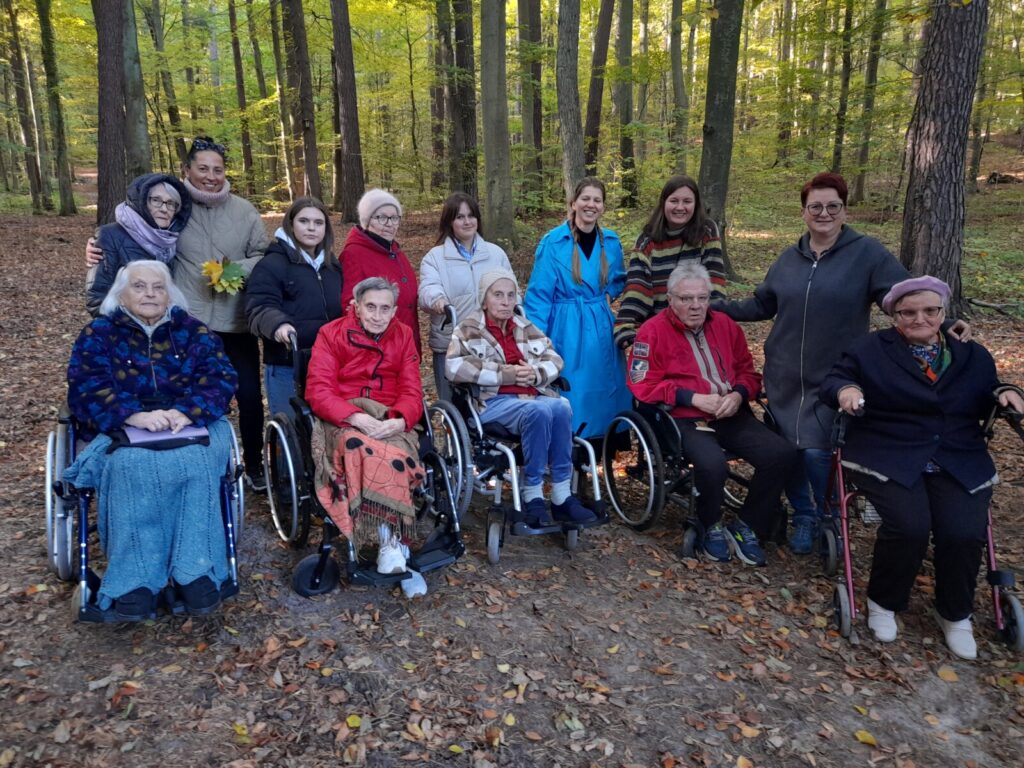 Las. Grupa seniorów na wózkach, wolontariuszy i opiekunów z DPS pozują do grupowego zdjęcia.