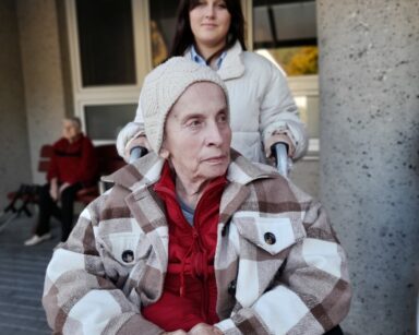 Zbliżenie. Młoda dziewczyna w jasnej kurtce pozuje do zdjęcia z seniorka siedzącą na wózku. Seniorka ubrana w bluzę i czapkę.